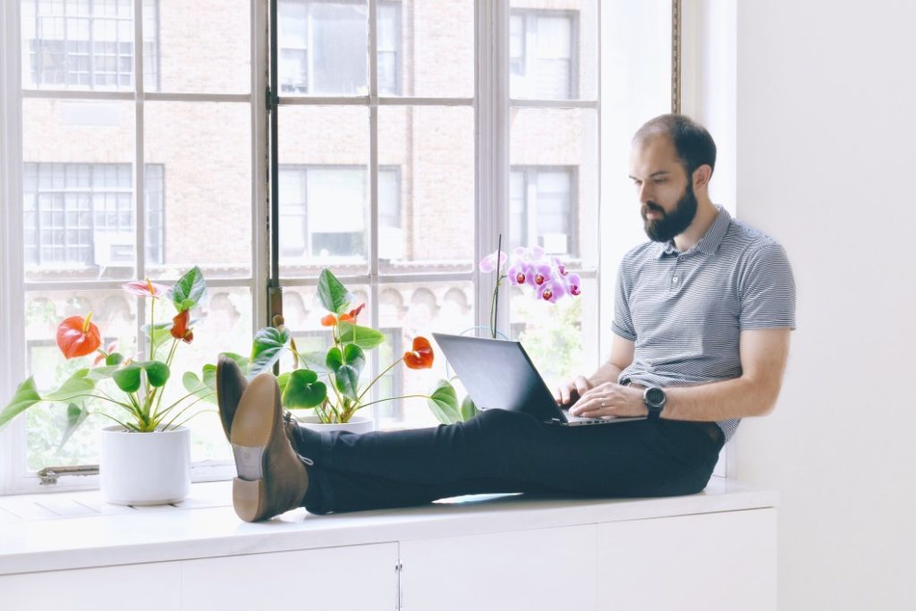 man sitting on window sill using computer by flowers