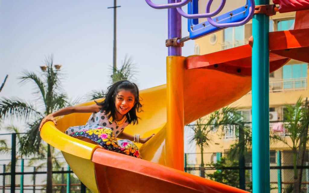 young girl in dress sliding down colorful slide by palm trees
