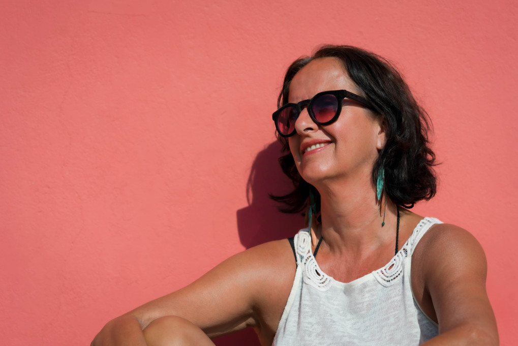 happy woman sitting by pink wall with sunglasses with