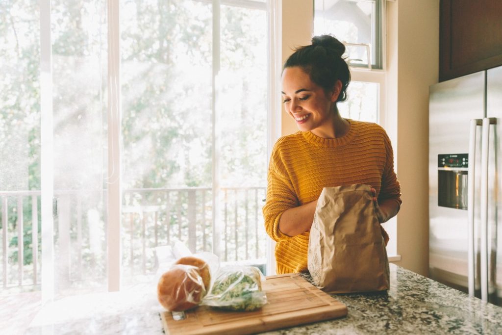 woman with brown bag smiling at groceries on the kitchen table