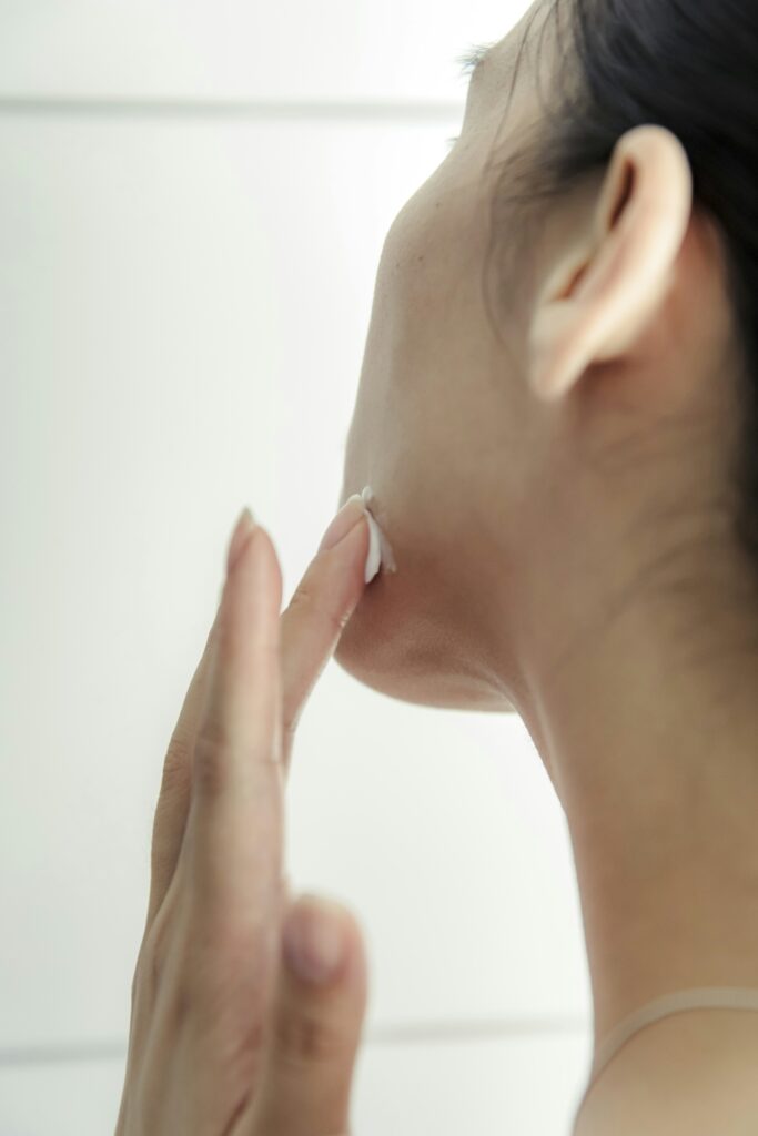 Back-left view of a woman’s head showing her ear and jawline as she applies moisturizer with elegantly extended fingers