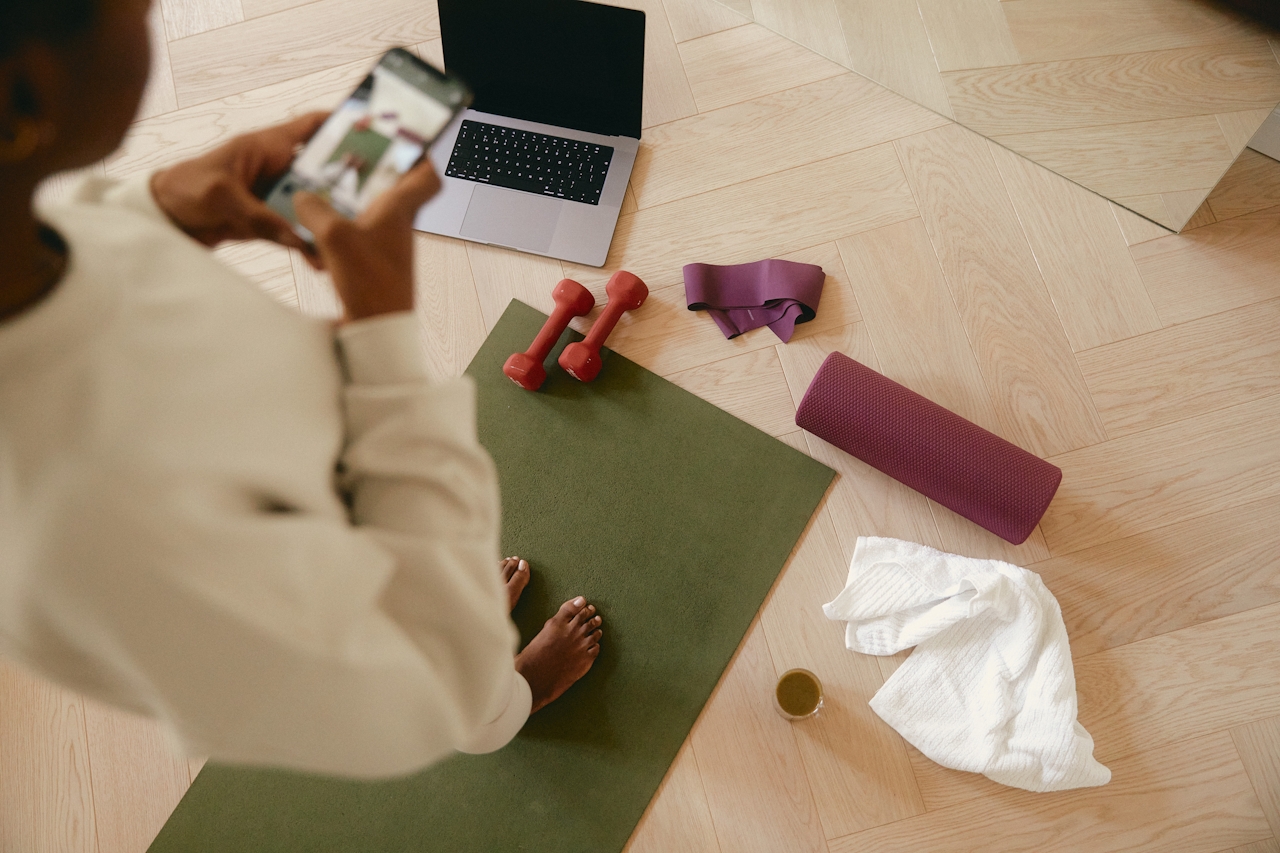 “Downward view from a woman’s shoulder of a yoga mat with a laptop, small red hand weights, a towel, and other workout gear arranged on it