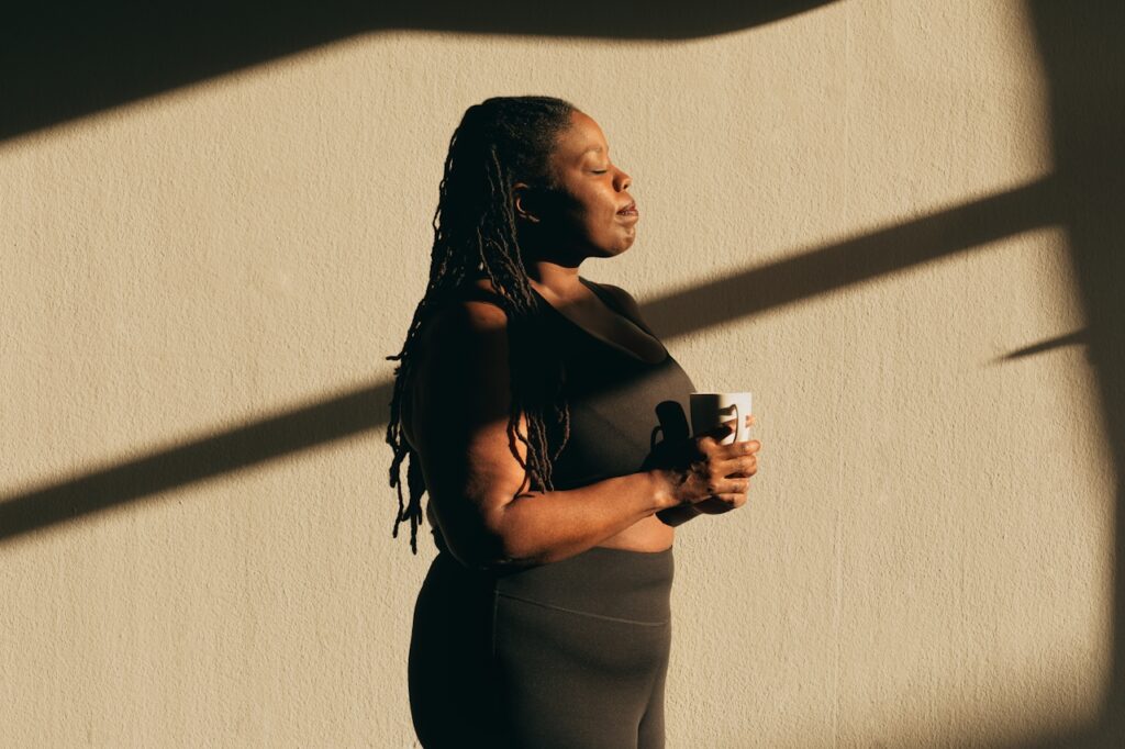 A woman standing in front of a concrete wall with sunlight streaming through a structure, eyes closed, holding a white mug, looking peaceful